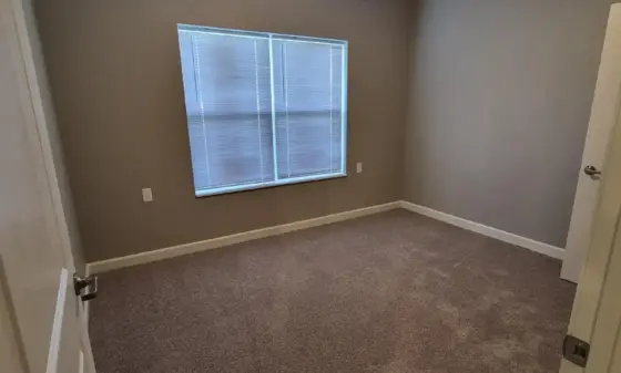 Small empty bedroom with beige walls, brown carpet, a window with blinds, and a ceiling fan with light overhead.