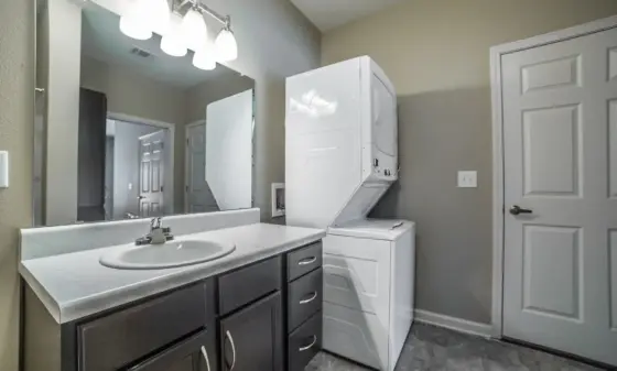 Laundry room with a stacked white washer and dryer beside a dark vanity and a large mirror, beige walls, and a three-bulb light fixture above.