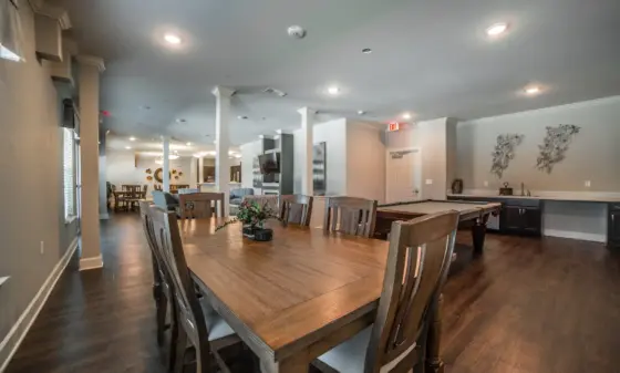 Dining area with a long wooden table and chairs in the foreground, ceiling lights, and a pool table visible in the background.