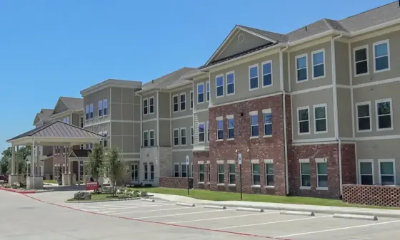 Exterior view of a multi-story apartment building with brick lower level and beige siding, white trim, and a covered entrance, under a clear blue sky; a spacious parking lot in the foreground.