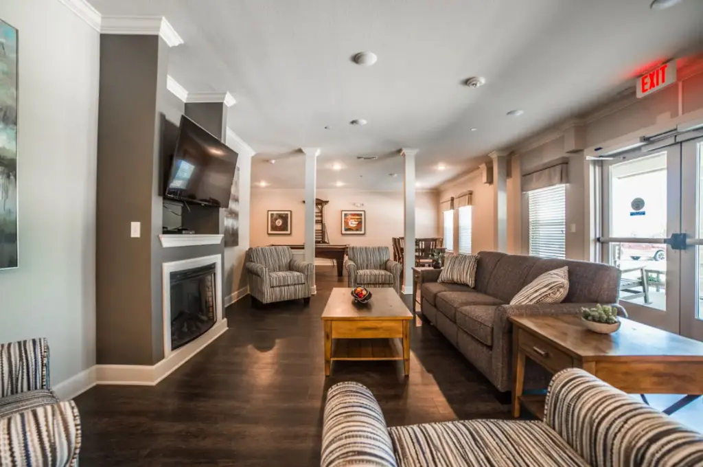 Lounge area with gray sofa, striped chairs, two wooden coffee tables, fireplace, and a wall-mounted TV; large windows on the right.