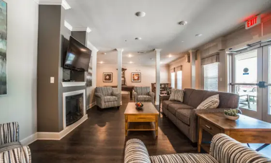 Lounge area with gray sofa, striped chairs, two wooden coffee tables, fireplace, and a wall-mounted TV; large windows on the right.