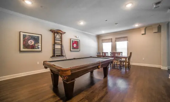 Basement game room with a large pool table in the foreground, billiards rack, framed pool-ball artwork, and a dining table by windows.