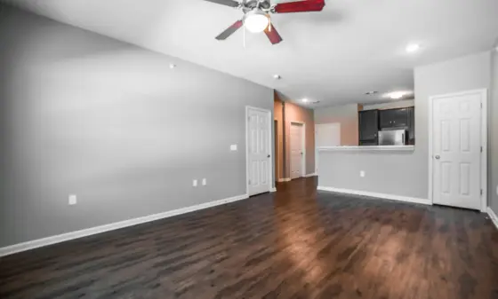 Bright, empty open-concept living room with gray walls, dark wood floors, and a ceiling fan; view into a kitchen with white cabinets and stainless appliances in the background.