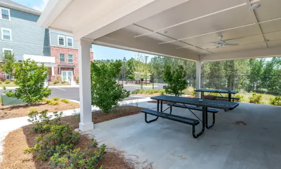 Covered outdoor seating area with two dark green metal picnic tables under a white ceiling; landscaped courtyard and blue-siding apartment building in the background.