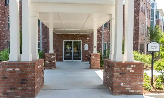 Covered entrance to a brick residential building with white columns and glass doors; a 'Resident Drop Off' sign is visible to the right.