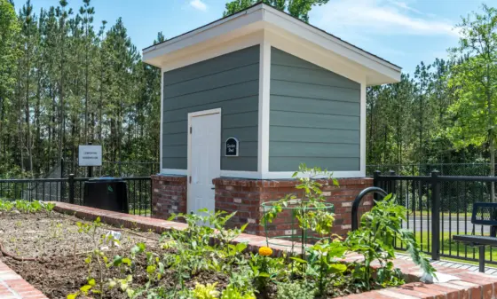 Small gray garden shed with white trim and brick foundation, set in a fenced garden with young plants and mulch beds.