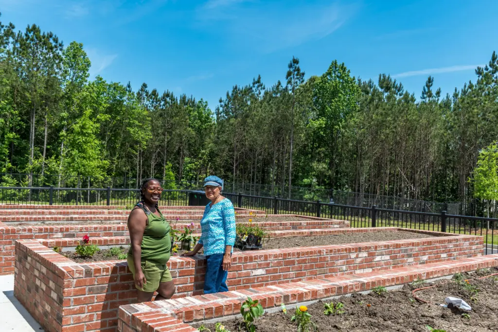 Two women stand on brick terraced garden beds with small flowering plants, smiling outdoors under a blue sky.