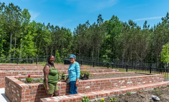 Two women stand on brick terraced garden beds with small flowering plants, smiling outdoors under a blue sky.