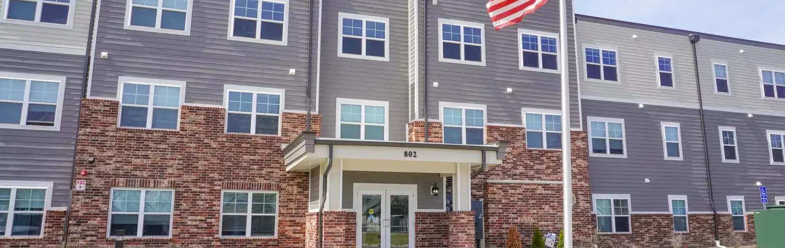 Apartment building with brick base, gray siding, and an American flag on a flagpole in front.] , hmm remove extra comma.