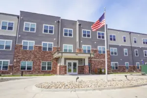 Apartment building with brick base, gray siding, and an American flag on a flagpole in front.] , hmm remove extra comma.