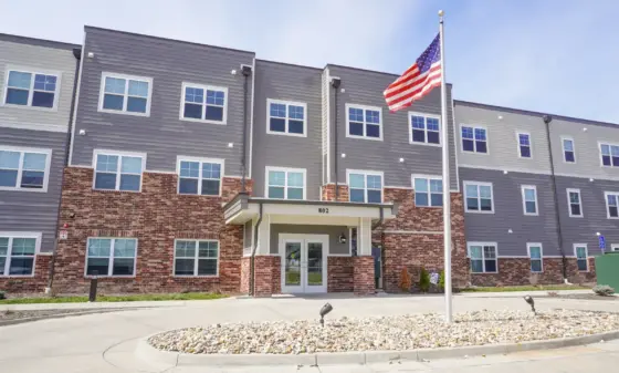 Apartment building with brick base, gray siding, and an American flag on a flagpole in front.] , hmm remove extra comma.