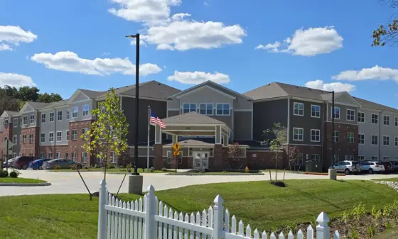 Front view of a modern multi-story apartment building with brick and gray siding, a covered entrance, and an American flag near the doorway under a blue sky.