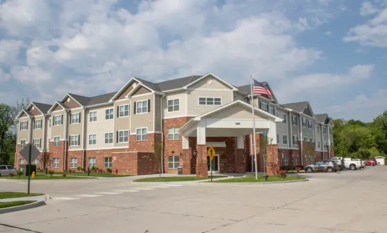 Three-story apartment building with beige siding and brick base under a blue sky with fluffy clouds; US flag on a pole near the entrance.