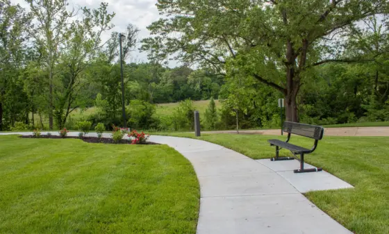 Curved concrete path through a green park, with a black bench and flower beds along the edge and trees in the background.