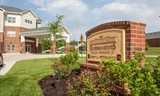 Entrance area of Riverbend Estates senior living community with brick buildings, a porte-cochère, and manicured landscaping under a blue sky. Sign: Riverbend Estates Senior Community II, brick pillars visible.