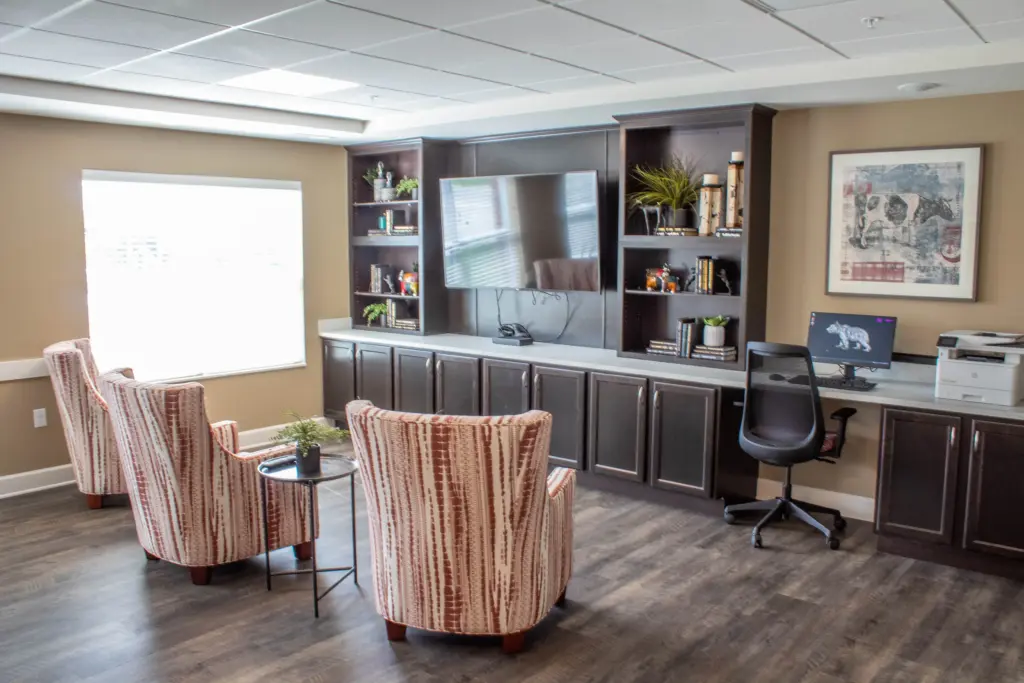 Modern home office lounge with dark wood built-ins, a wall-mounted TV, shelves, and a desk with computer and printer nearby. Striped upholstered chairs surround a small glass table.