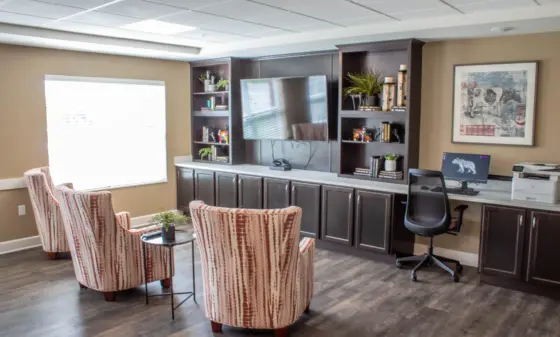 Modern home office lounge with dark wood built-ins, a wall-mounted TV, shelves, and a desk with computer and printer nearby. Striped upholstered chairs surround a small glass table.