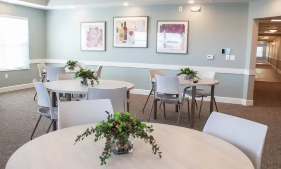 Bright dining area with round white tables, gray chairs, and a vase of greenery in the center; wall art and soft blue walls.
