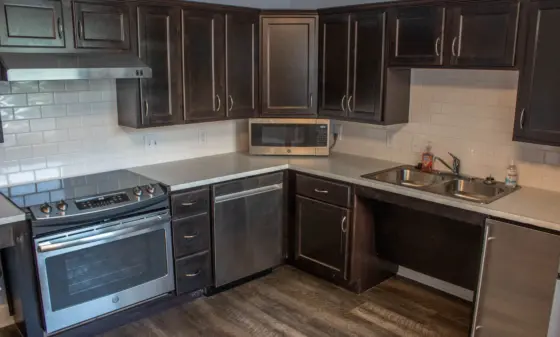 Dark wood kitchen with stainless steel stove, microwave, and double sink along white subway tile backsplash and gray countertops.