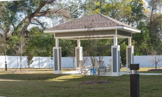 Gazebo in a park with white posts and a shaded seating area, several rocking chairs nearby.