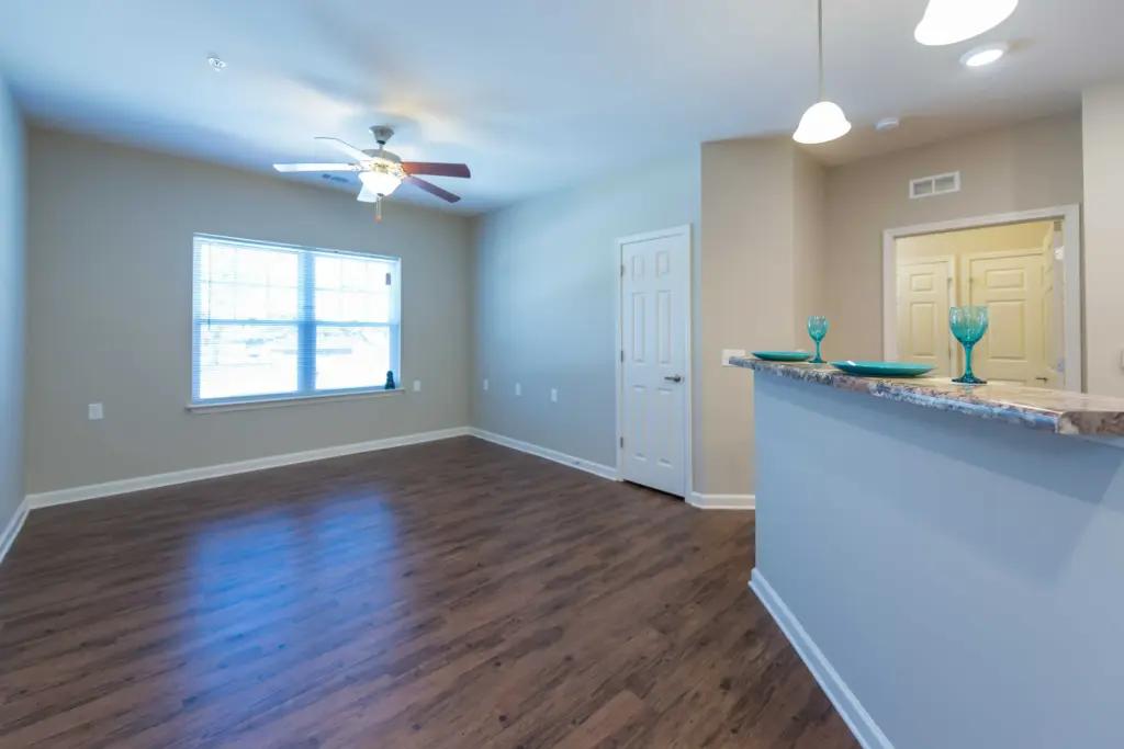 Open living area with wood floors, beige walls, a ceiling fan, large window, and a kitchen counter displaying turquoise glassware.