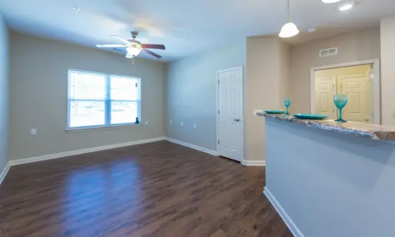 Open living area with wood floors, beige walls, a ceiling fan, large window, and a kitchen counter displaying turquoise glassware.