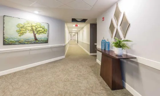Wide hotel hallway with a landscape painting of a tree on the left and diamond-shaped mirrors above a wooden console on the right, leading to an exit in the distance.