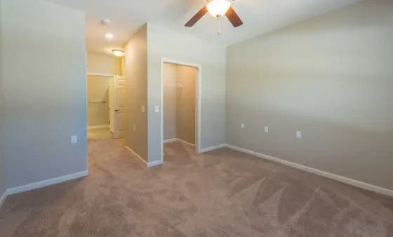 Empty beige bedroom with carpet, ceiling fan, and multiple doorways leading to a closet and hallway.