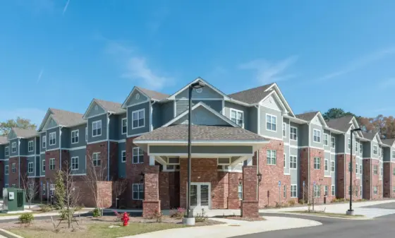 Modern apartment building with gray-blue siding and brick base under a clear blue sky; multiple gabled roofs and white trim.