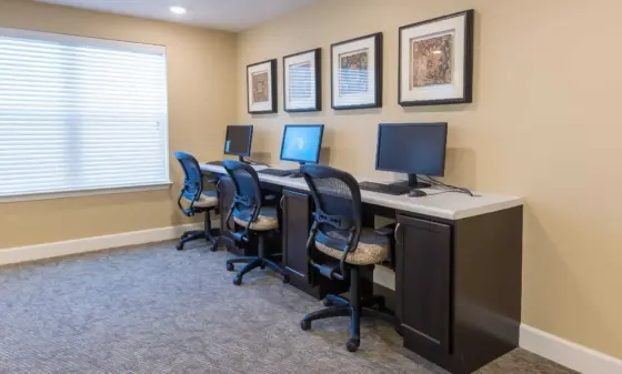 Three computer desks with monitors and chairs against a beige wall, with framed art above and a window with blinds to the left.