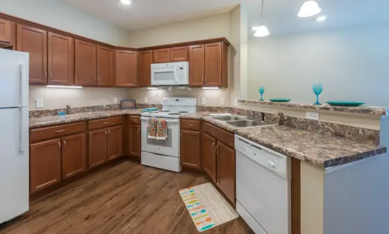 U-shaped kitchen with brown wooden cabinets, white appliances (fridge, stove, microwave), and granite countertops, plus a breakfast bar with turquoise glassware on top.