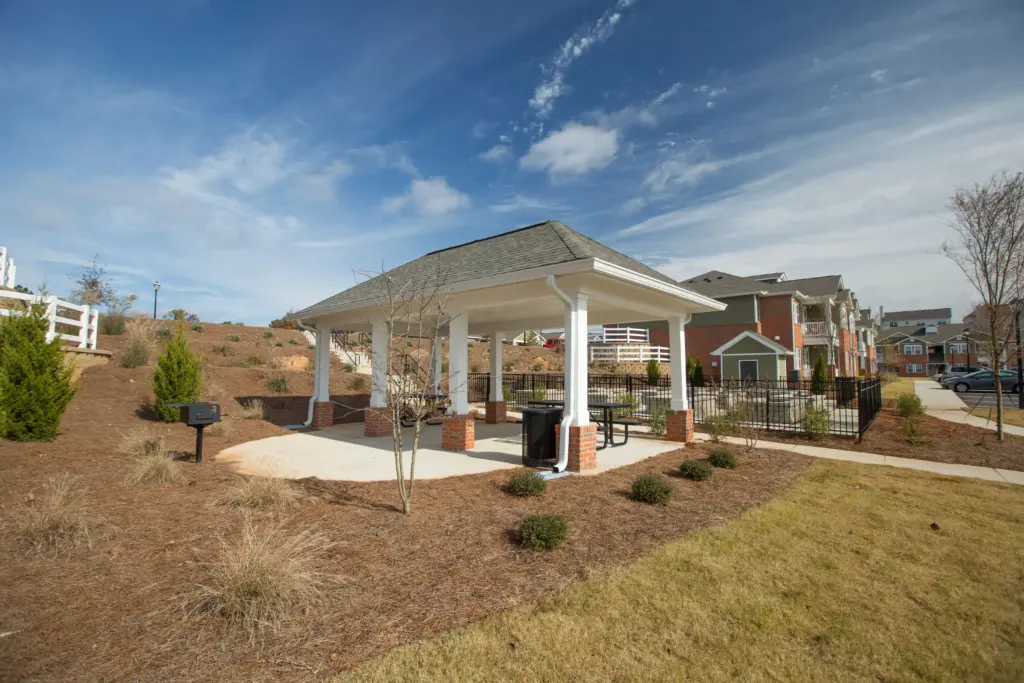 White-pigtail pavilion with brick columns and picnic tables in a landscaped, suburban apartment complex under a bright blue sky with scattered clouds