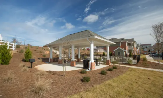 White-pigtail pavilion with brick columns and picnic tables in a landscaped, suburban apartment complex under a bright blue sky with scattered clouds
