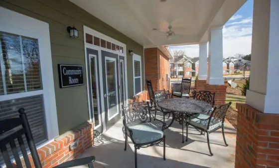 Covered porch with ornate black metal patio furniture and a round table; a 'Community Room' sign is mounted on green siding near glass doors, brick columns in view, neighborhood houses in background.