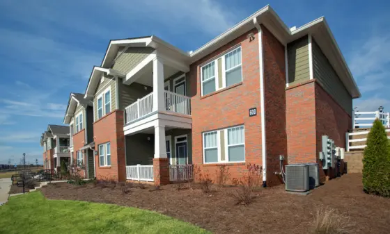 Brick apartment building with white railings and balconies under a clear blue sky. Frontal view shows multiple windows and entryways on a sunny day.