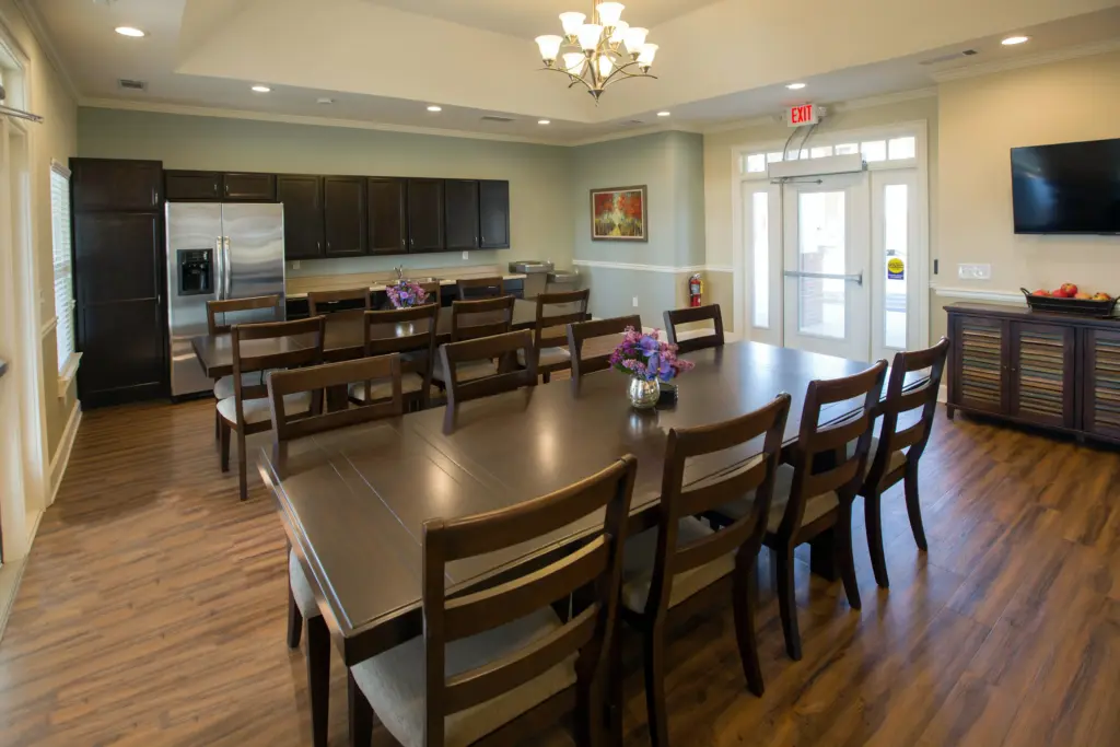 Roomy dining/meeting area with a long dark wood table and many chairs, a kitchen line of dark cabinets and a stainless fridge, and a chandelier overhead.