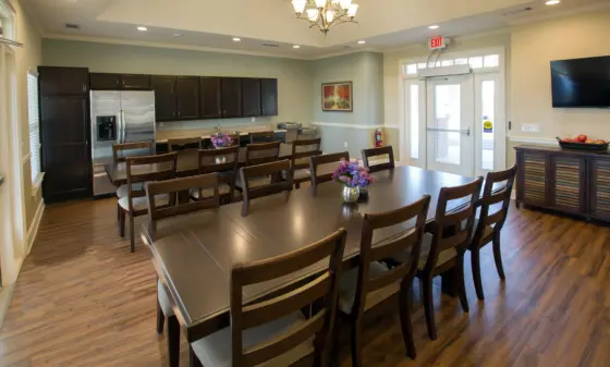 Roomy dining/meeting area with a long dark wood table and many chairs, a kitchen line of dark cabinets and a stainless fridge, and a chandelier overhead.
