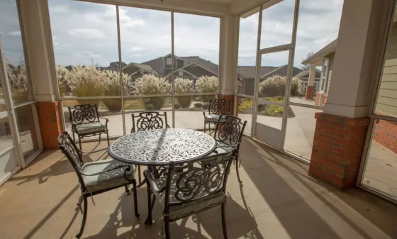 Screened porch with round ornate metal table and matching chairs, overlooking a yard with tall ornamental grasses and nearby houses in the distance.