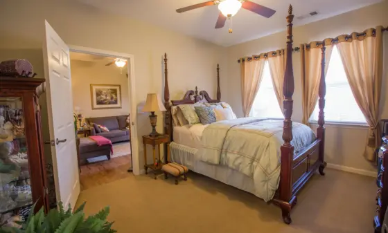 Bedroom with a dark wood four-poster bed, beige bedding, and cream curtains; open doorway to a living room on the left.