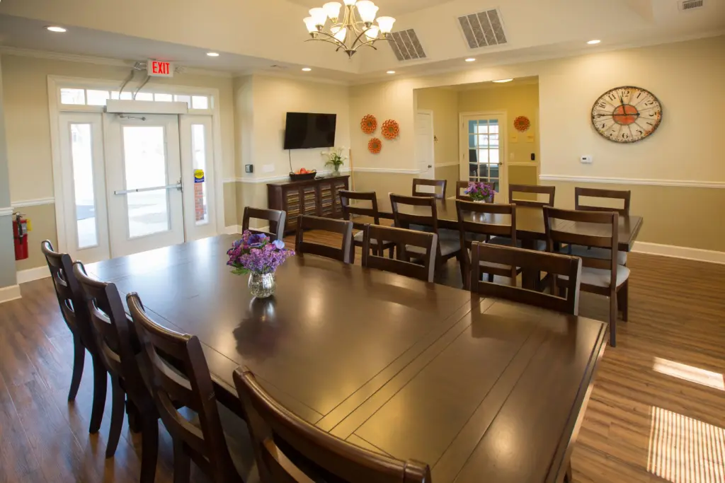 Communal dining room with a long dark wooden table, chairs, and purple flowers in glass vases, beige walls, and a large round wall clock.”