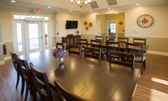 Communal dining room with a long dark wooden table, chairs, and purple flowers in glass vases, beige walls, and a large round wall clock.”