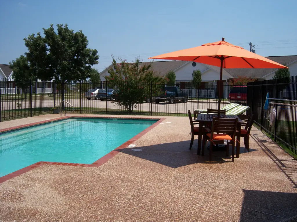 Poolside patio with an orange umbrella, wooden table and chairs, and a fenced yard in a suburban setting under a clear blue sky.