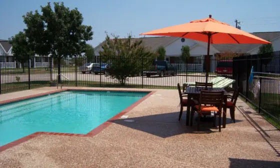 Poolside patio with an orange umbrella, wooden table and chairs, and a fenced yard in a suburban setting under a clear blue sky.