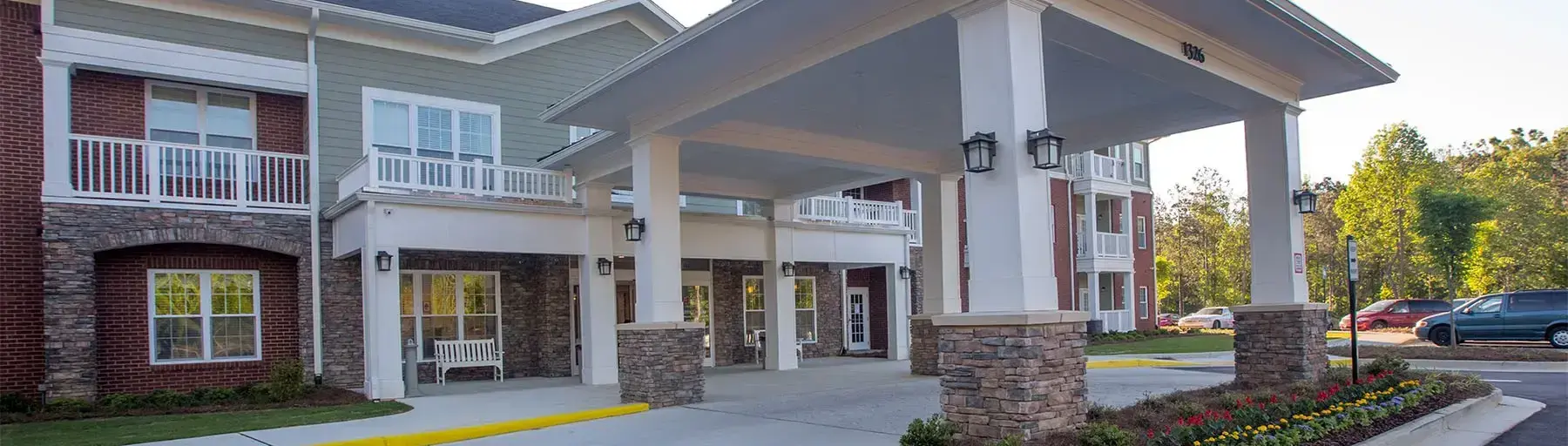 Porte-cochère entrance to a multiunit building with stone pillars, white columns, brick and siding exterior, and landscaped flower beds nearby.