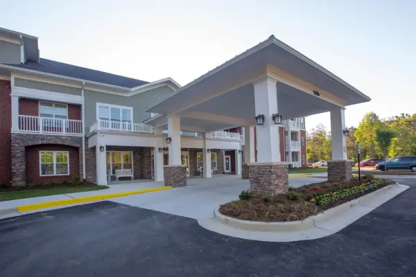 Porte-cochère entrance to a multiunit building with stone pillars, white columns, brick and siding exterior, and landscaped flower beds nearby.
